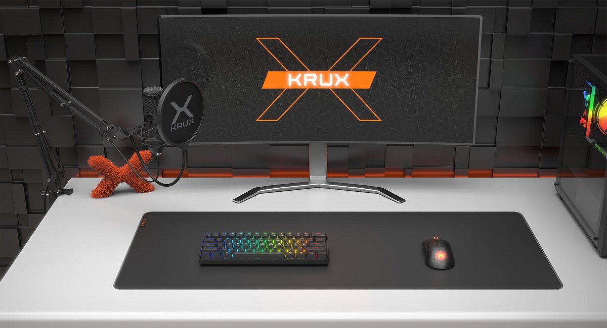 Na biurku znajduje się monitor z logo \'KRUX\', klawiatura z podświetleniem RGB, mysz oraz mikrofon na statywie z pop filtrem. W tle widać komputer z podświetleniem RGB.