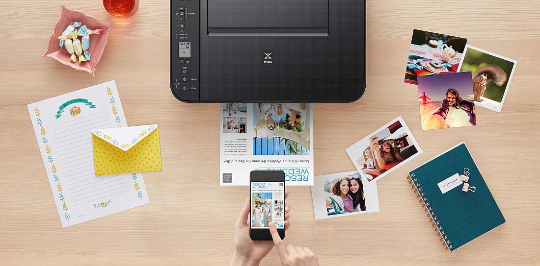 Na biurku znajduje się urządzenie wielofunkcyjne z widocznym logo Pixma, obok którego leży telefon komórkowy z wyświetlonym zdjęciem. Wokół rozłożone są kolorowe zdjęcia, notes, koperta oraz kartka papieru z kolorowym wzorem.