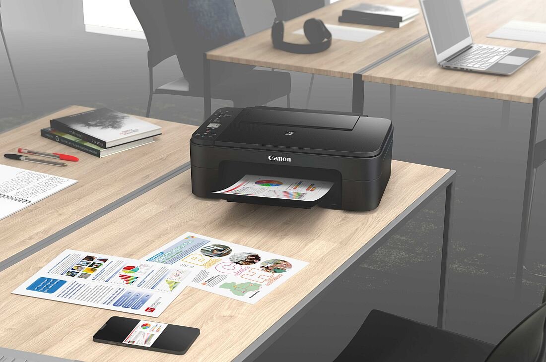 Na drewnianym biurku znajduje się czarne urządzenie wielofunkcyjne z logo Canon, z którego wysuwa się kolorowy wydruk. W tle widać inne biurka z laptopem, książkami i słuchawkami.