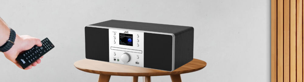 Wieża JVC RD-E874B z radiem internetowym, DAB+ i CD, wieża stojąca po skosie na stoliku, ręka trzymająca pilota