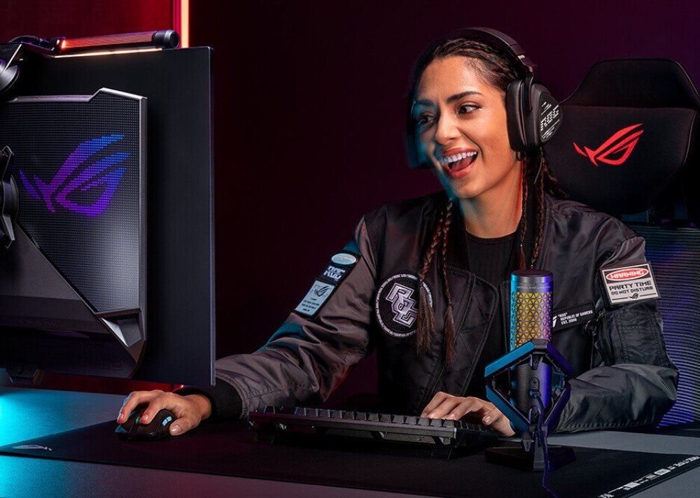 Monitor ASUS ROG Strix XG27AQDPG Uśmiechnięta kobieta w słuchawkach siedząca przed monitorem gamingowym w ciemnym pomieszczeniu, na biurku znajduje się klawiatura, mysz oraz profesjonalny mikrofon, funkcje, redukcja powidoków, ochrona wzroku
