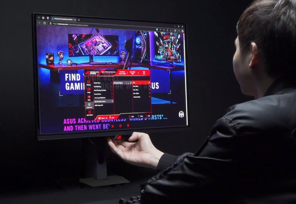 Monitor ASUS ROG Strix XG27AQDPG Zbliżenie na dłoń użytkownika obsługującą przyciski sterujące pod dolną krawędzią monitora, na ekranie wyświetlone jest menu ustawień systemowych 'OLED Care' na tle strony internetowej, ochrona matrycy, czujnik obecności, funkcje