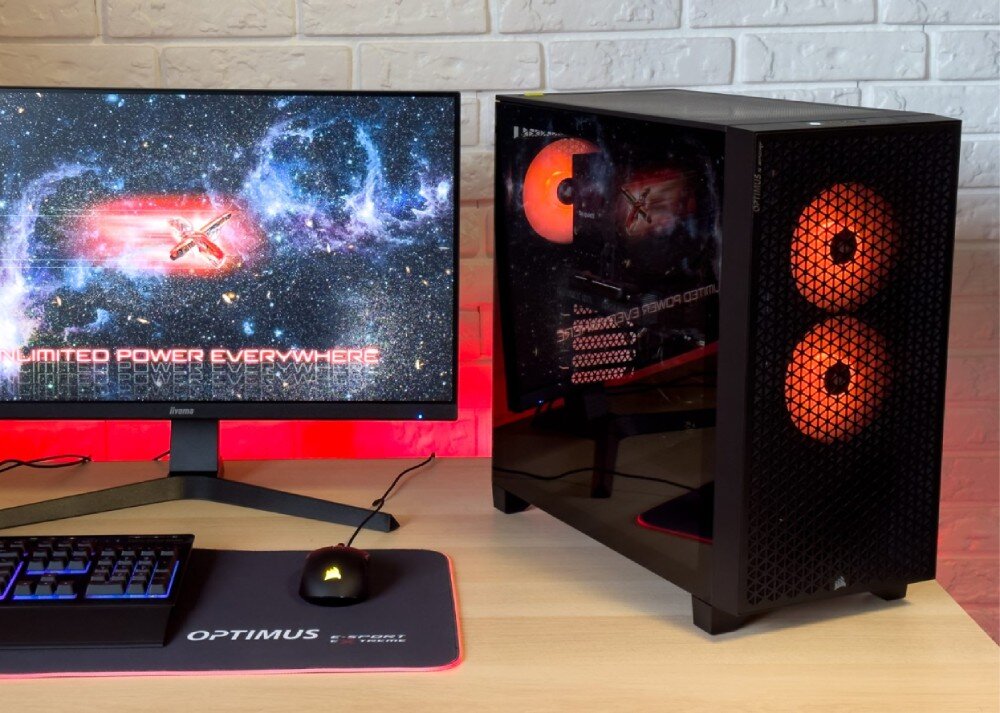 Na biurku znajduje się monitor z wyświetlonym obrazem kosmosu i tekstem \'UNLIMITED POWER EVERYWHERE\', obok stoi komputer z podświetleniem wentylatorów na pomarańczowo. Na podkładce pod mysz widnieje napis \'OPTIMUS\'.