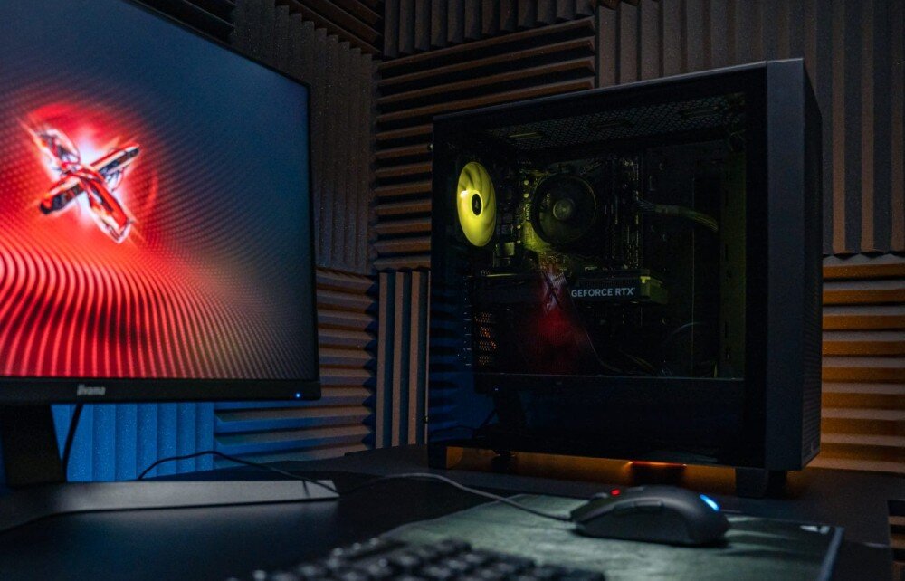 Komputer OPTIMUS E-Sport GA520T-CR10 Komputer stacjonarny w czarnej obudowie z przeszklonym bokiem stoi na biurku obok monitora wyświetlającego czerwoną grafikę. Wewnątrz obudowy widoczny jest świecący na żółto wentylator, opis