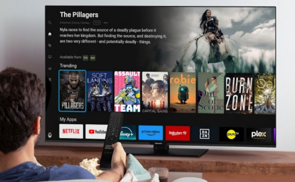 Telewizor PANASONIC TB-65W61AEZ Smart TV TiVo sterowanie głosowe, osoba siedząca na kanapie przed telewizorem, trzymająca pilot w ręku