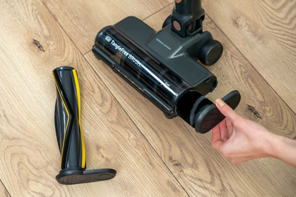 Na drewnianej podłodze znajduje się odkurzacz z widoczną szczotką oznaczoną jako \'TangleFree BRUSH\'. Obok leży wyjęty element szczotki, a ręka trzyma okrągłą część odkurzacza.