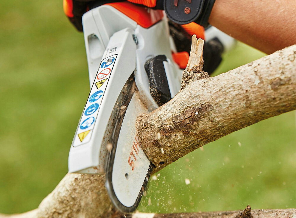 Piła akumulatorowa STIHL GTA 26 ogród osoba tnie gałąź w tle trawa asymetryczna ruchoma osłona zabezpiecza łańcuch w zakresie 90 stopni poprawiając widoczność ciętego elementu ergonomiczny uchwyt pewność chwyt większy komfort i bezpieczeństwo pracy