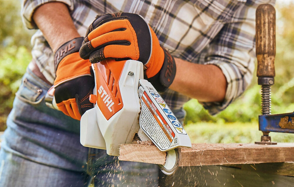 Piła akumulatorowa STIHL GTA 26 ogród osoba tnie deskę w tle krzewy funkcja bezpiecznego rozruchu przycisk odblokowania ochrona przed przypadkowym uruchomieniem sygnalizacja LED informuje o aktywacji pięciosekundowy limit na rozpoczęcie pracy bezpieczeństwo obsługi