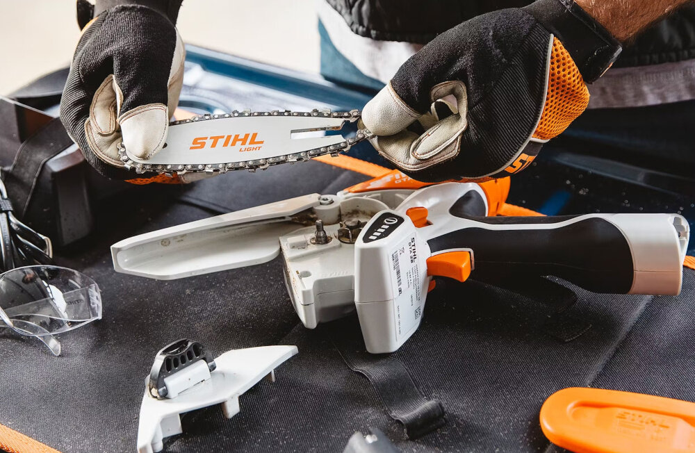 Piła akumulatorowa STIHL GTA 26 blat stołu osoba trzyma łańcuch w dłoniach na stole leży piła beznarzędziowy system wymiany i napinania łańcucha ręczne smarowanie umożliwia precyzyjne dozowanie oleju dłuższa żywotność elementów tnących