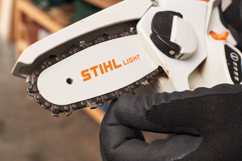 Na zdjęciu widoczna jest prowadnica piły z łańcuchem, na której znajduje się napis \'STIHL LIGHT\'. Osoba w rękawicy trzyma piłę, a w tle widać rozmyte elementy wnętrza.