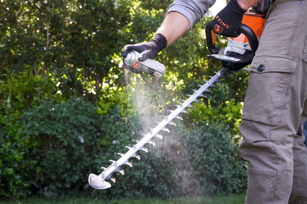 Osoba w rękawicach trzyma nożyce do żywopłotu i spryskuje je aerozolem. Na puszce widoczne jest logo STIHL.