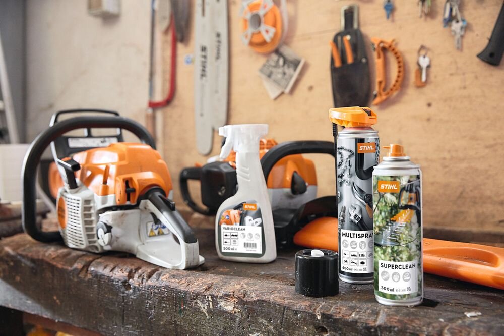 Na drewnianym stole znajdują się piły łańcuchowe oraz produkty czyszczące marki STIHL, w tym Multispray 400 ml. W tle widać narzędzia wiszące na ścianie.