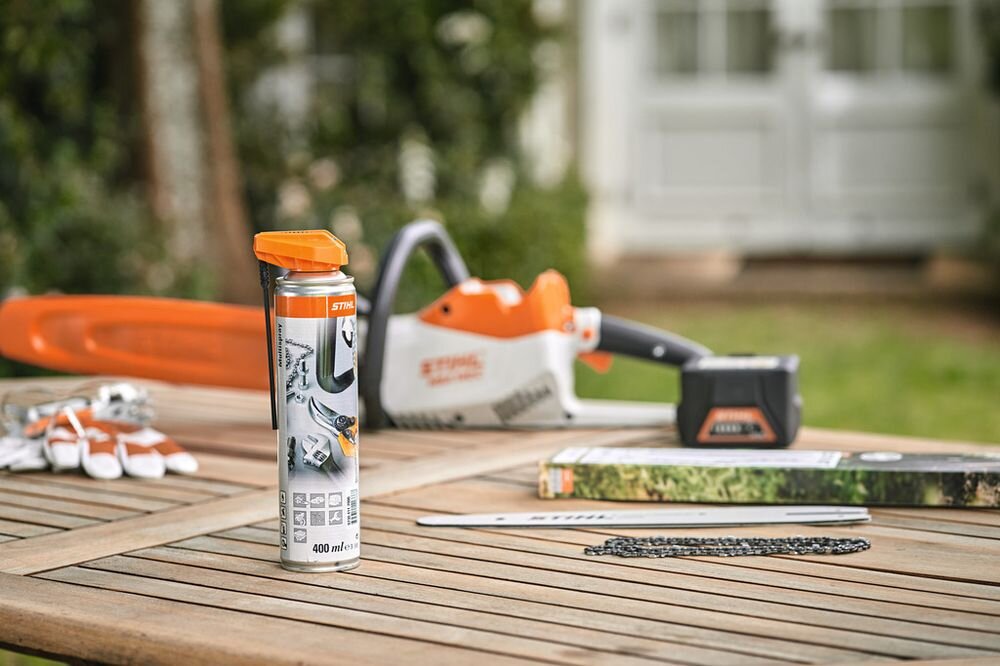 Na drewnianym stole znajduje się puszka uniwersalnego środka czyszczącego STIHL Multispray 400 ml. W tle widoczna jest piła łańcuchowa oraz akcesoria do jej obsługi.