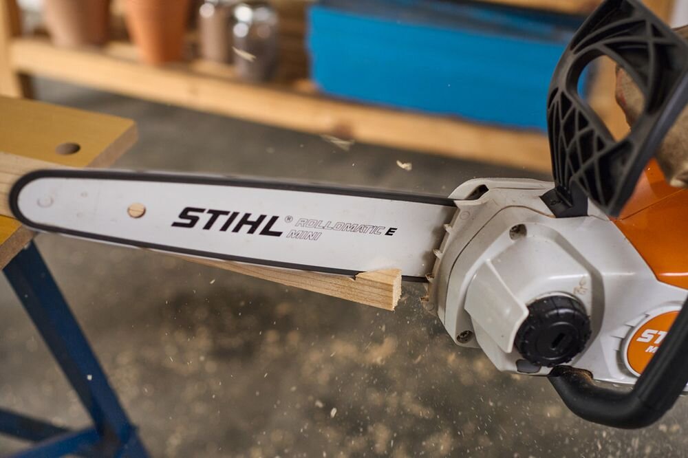 Na zdjęciu widać piłę łańcuchową z widocznym napisem \'STIHL ROLLOMATIC E MINI\'. Piła przecina kawałek drewna, a w tle znajdują się rozmyte elementy warsztatu.