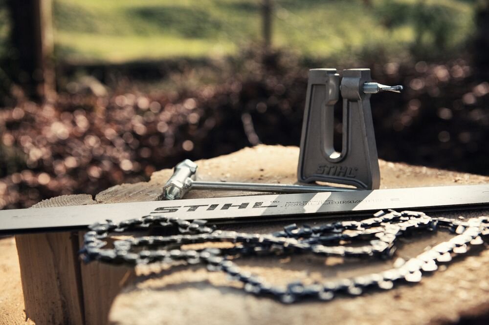 Na drewnianym pniu leży łańcuch do piły oraz prowadnica z widocznym napisem \'STIHL\'. W tle znajduje się narzędzie do ostrzenia z logo \'STIHL\'.
