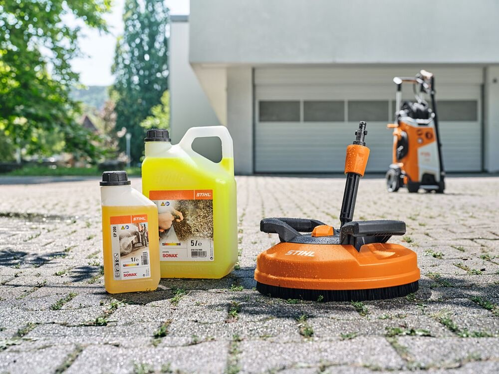 Na obrazie widoczne są trzy produkty marki STIHL: dwa pojemniki z płynem czyszczącym oraz pomarańczowe urządzenie czyszczące. W tle znajduje się budynek z zamkniętymi drzwiami garażowymi oraz sprzęt czyszczący na kółkach.