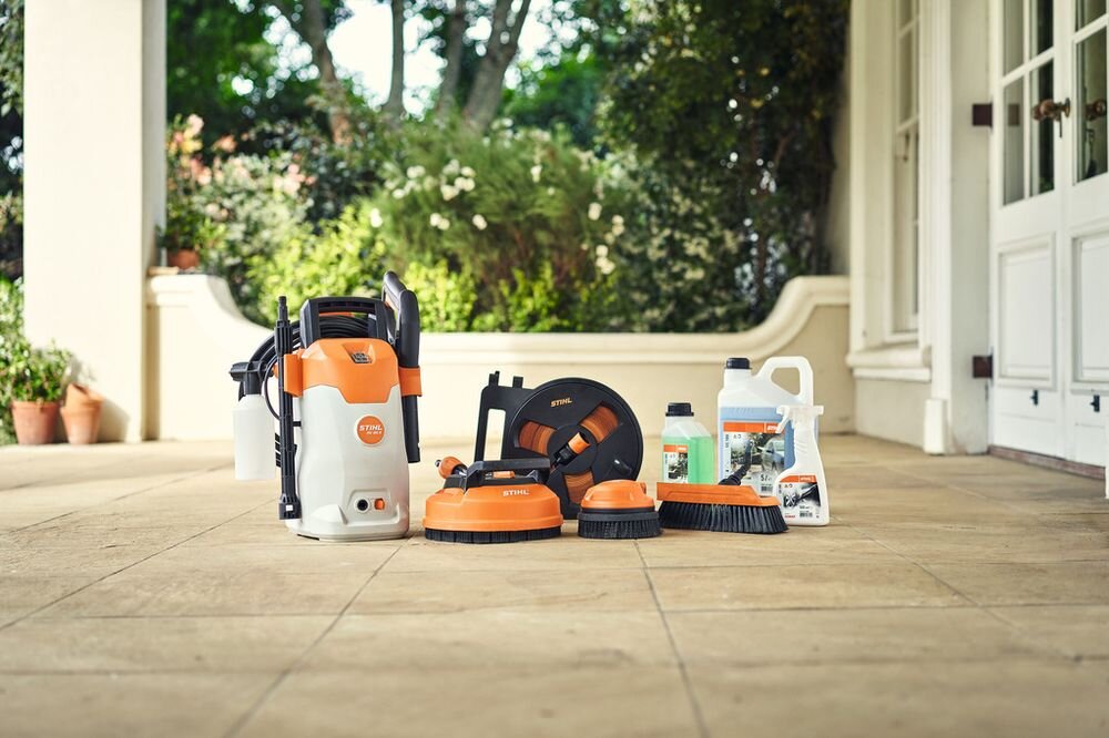 Na tarasie znajduje się zestaw narzędzi czyszczących w kolorach pomarańczowym i białym, z widocznym logo STIHL. W tle widać rośliny i białe drzwi.