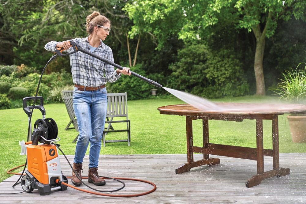 Osoba używa myjki ciśnieniowej do czyszczenia drewnianego stołu na tarasie. Na urządzeniu widoczne jest logo \'STIHL\'.
