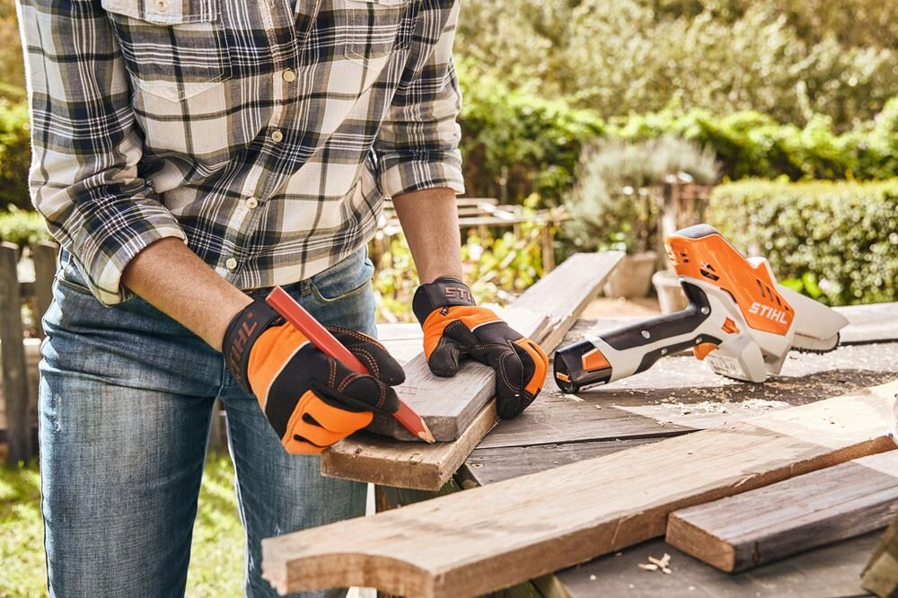 Osoba w koszuli w kratę i dżinsach pracuje z drewnem, używając pomarańczowo-czarnych rękawic roboczych z logo STIHL. Na stole obok znajduje się narzędzie z widocznym napisem STIHL.