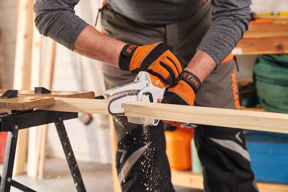 Osoba w pomarańczowych rękawicach roboczych z logo STIHL używa piły łańcuchowej do cięcia drewnianej deski. W tle widoczne są narzędzia i materiały budowlane.
