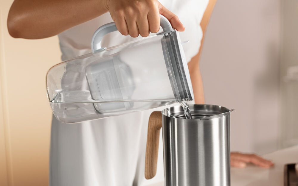 Osoba nalewa wodę z przezroczystego dzbanka do metalowego naczynia. Dzbanek ma biały uchwyt i widoczny jest w nim filtr.