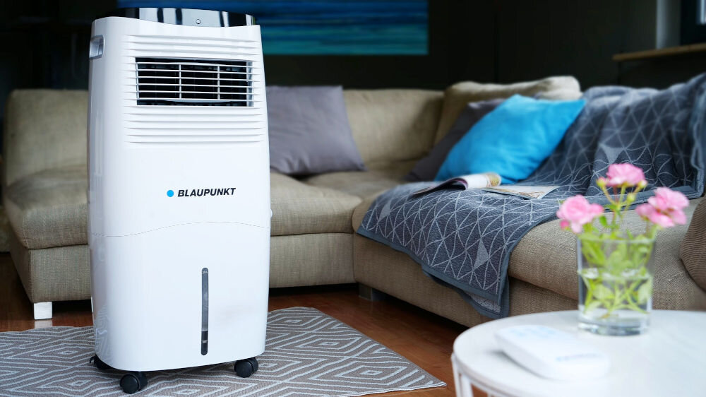 Na zdjęciu znajduje się biały klimator z logo BLAUPUNKT, ustawiony na dywanie w salonie z beżową sofą i poduszkami w tle. Na stoliku obok widać szklany wazon z różowymi kwiatami.