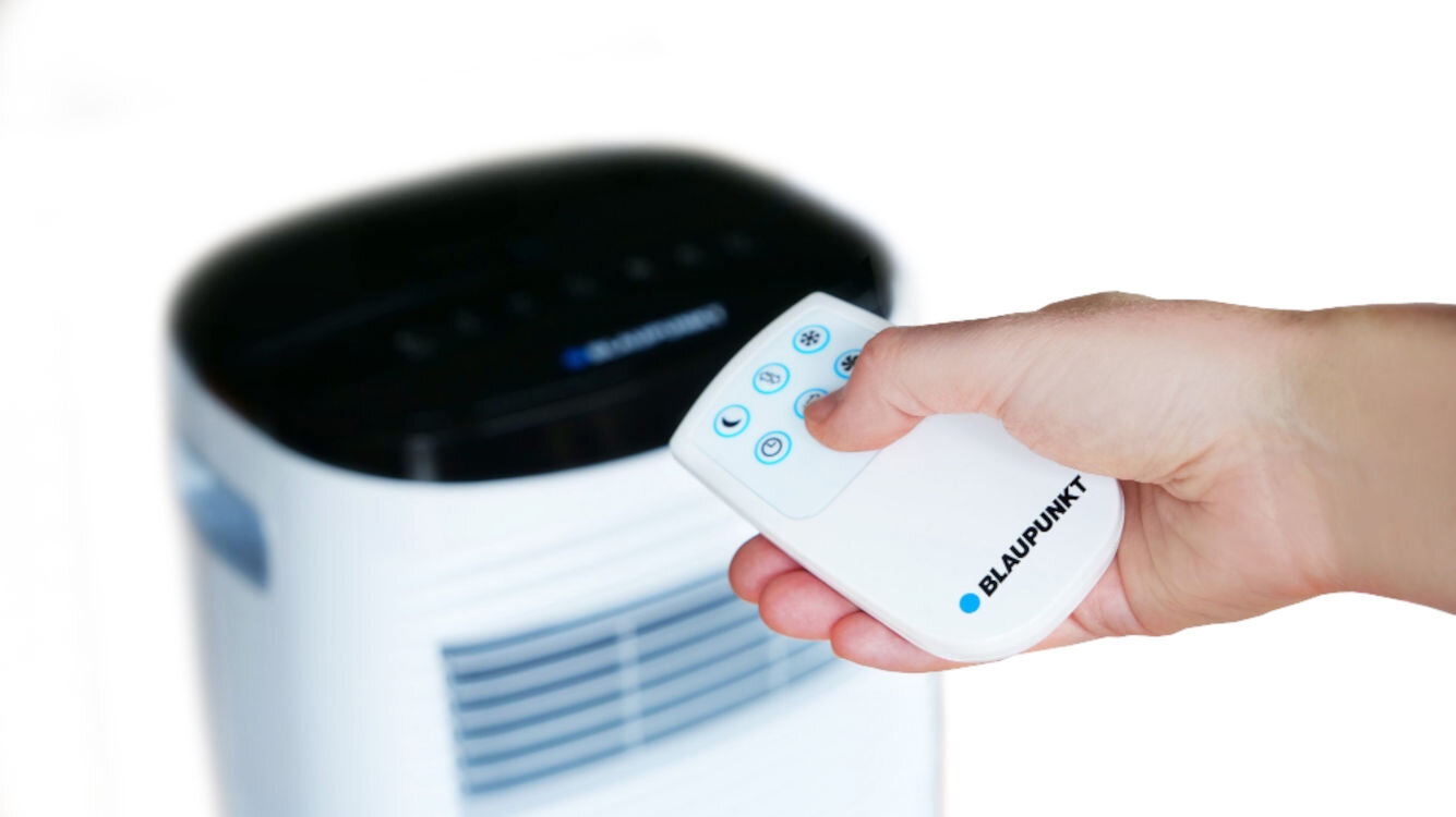 Osoba trzyma pilot z napisem \'BLAUPUNKT\' skierowany w stronę klimatyzatora. Klimatyzator ma czarny panel górny i widoczne kratki wentylacyjne.