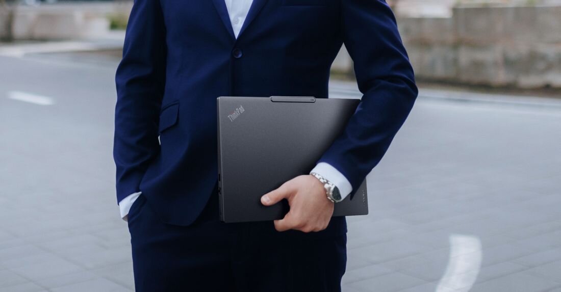 Osoba trzyma zamknięty laptop Lenovo ThinkPad E14 Gen 7 w kolorze Thunder Black stojąc na zewnątrz w przestrzeni miejskiej - Design i kolorystyka — Thunder Black