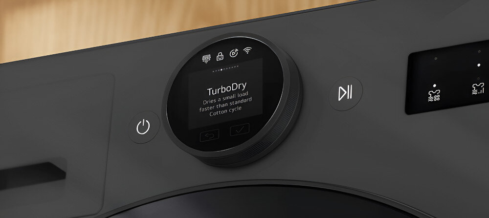 SUSZARKA LG RHX7010TBB Na zdjęciu pokazano zbliżenie na panel sterowania suszarki LG. Centralnie widoczny jest okrągły wyświetlacz z informacją o funkcji „TurboDry”, a po bokach umieszczone są przyciski start/pauza oraz zasilania. Widoczne są również ikony dotykowe i minimalistyczny design panelu