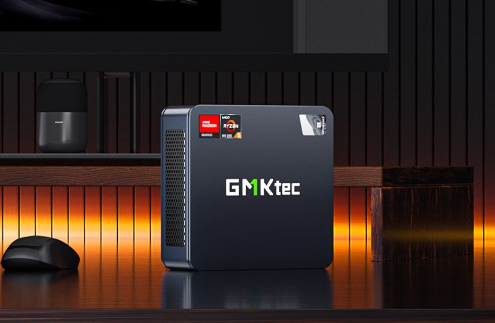 Komputer GMKTEC M6 Małe pudełkowe urządzenie z logo 'GMKtec' ustawione pionowo na biurku, obok myszy i monitora, w tle ciemne, podświetlone tło, opis