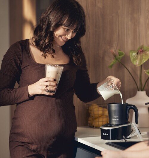 Osoba wlewa mleko do spieniacza marki Amica, trzymając w drugiej ręce szklankę z napojem. Na urządzeniu widoczne jest logo \'Amica\'.