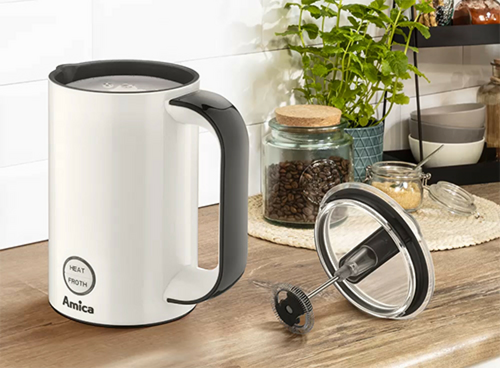 Na blacie kuchennym znajduje się biały spieniacz do mleka z czarnym uchwytem i przyciskami oznaczonymi jako \'HEAT\' i \'FROTH\'. Obok leży pokrywka z mechanizmem spieniającym, a w tle widoczne są słoiki z kawą i rośliny.