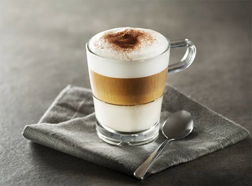 Szklanka z warstwową kawą i pianką na wierzchu stoi na szarej serwetce obok łyżeczki.