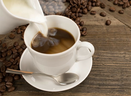 Na drewnianym stole stoi filiżanka z kawą, do której wlewane jest mleko z białego dzbanka. Obok filiżanki leży łyżeczka, a wokół rozsypane są ziarna kawy.