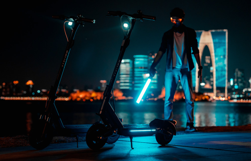 Hulajnoga elektryczna NAVEE GT3 45 km 350W Amortyzacja Czarny 10 cali miasto noc dwie hulajnogi stoją oparte na stopkach hulajnogi mają zapalone przednie światła w tle stoi osoba z mieczem świetlnym pełny zestaw świateł LED inteligentne rozwiązania reflektory z czujnikiem światła samoczynnie dostosowują natężenie do warunków otoczenia wbudowane kierunkowskazy sygnalizowanie manewrów