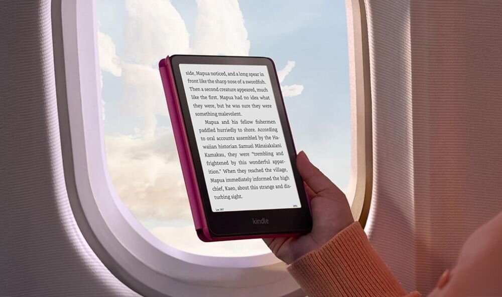 Osoba trzyma czytnik e-booków Kindle z widocznym tekstem na ekranie, siedząc przy oknie samolotu z widokiem na niebo i chmury.