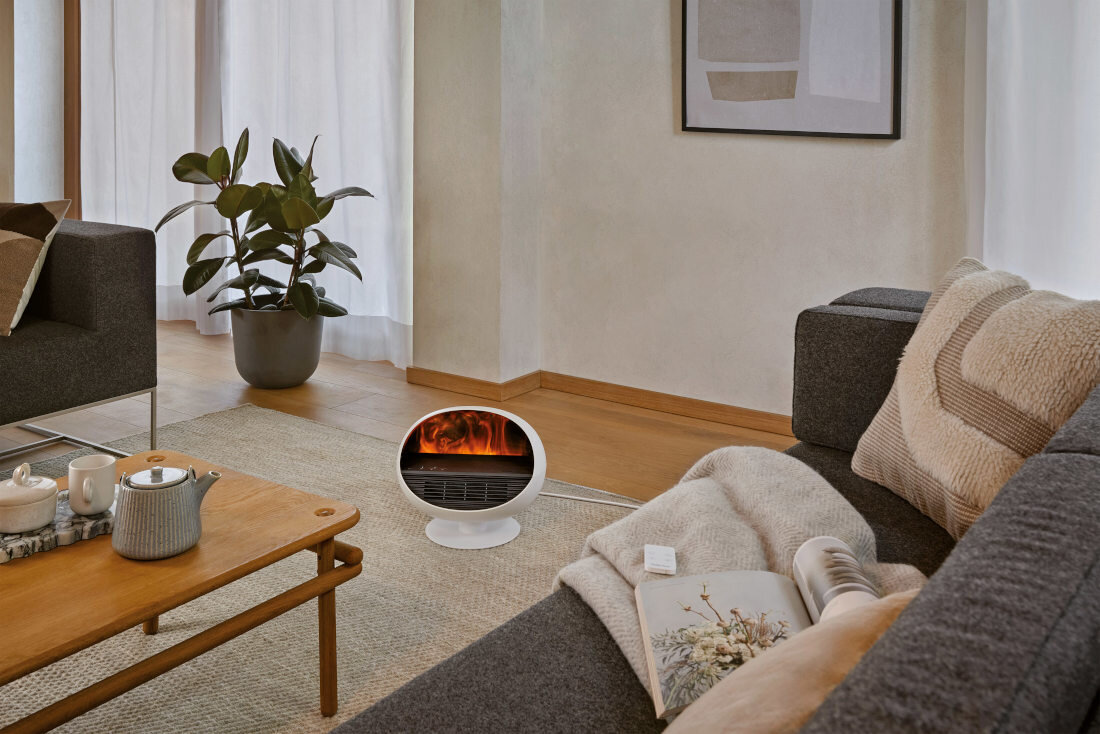 W salonie na dywanie stoi biały termowentylator z widocznym efektem płomieni. W tle znajduje się szara sofa, drewniany stolik z czajnikiem i filiżankami oraz roślina w doniczce.