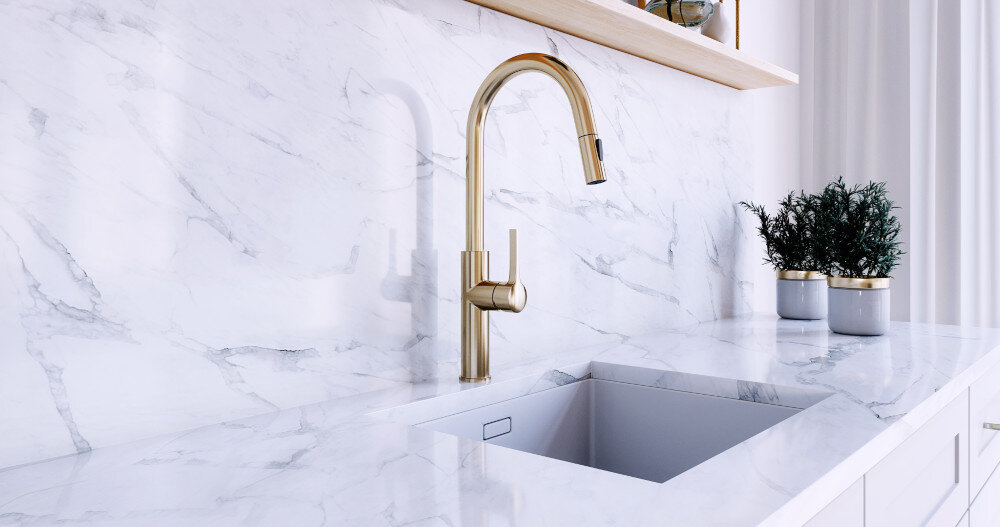 Złota matowa bateria kuchenna zamontowana na marmurowym blacie z wbudowanym zlewem. W tle widoczne są dwie doniczki z roślinami na blacie oraz półka z dekoracjami.