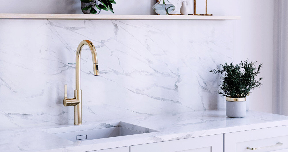 Na obrazie widoczna jest złota bateria zamontowana nad białym zlewem na tle marmurowej ściany. Na blacie stoi doniczka z rośliną, a na półce znajdują się dekoracje i rośliny.