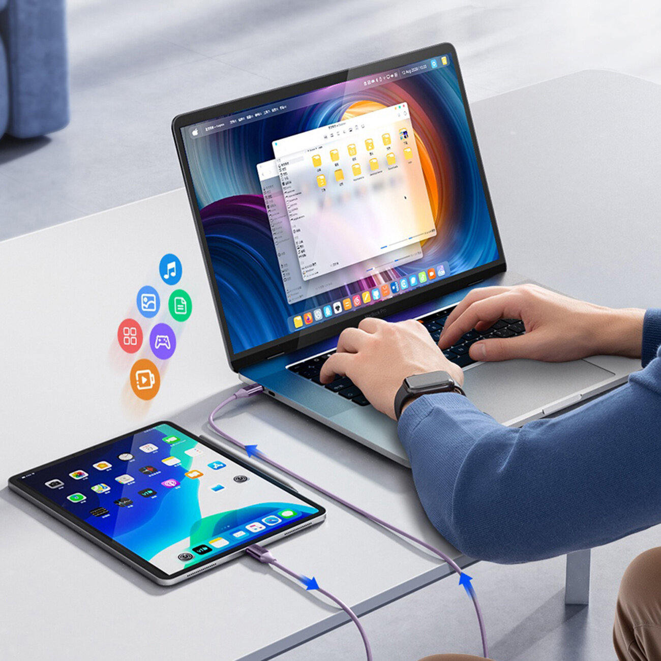 Osoba używa laptopa, do którego podłączony jest fioletowy kabel USB-C, łączący również tablet leżący na stole. Na ekranie laptopa widoczne są otwarte okna z folderami, a na tablecie ikony aplikacji.
