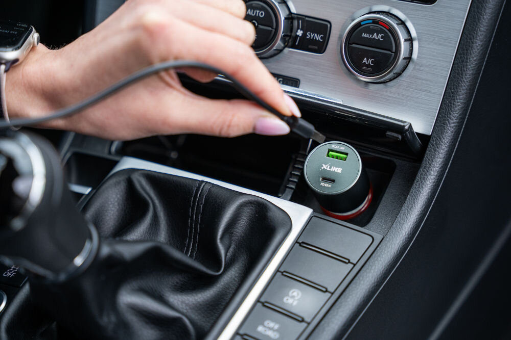 Osoba podłącza kabel do ładowarki samochodowej umieszczonej w gnieździe zapalniczki. Na ładowarce widoczne jest logo \'XLINE\' oraz napisy \'QC3.0\' i \'PD65W\'.