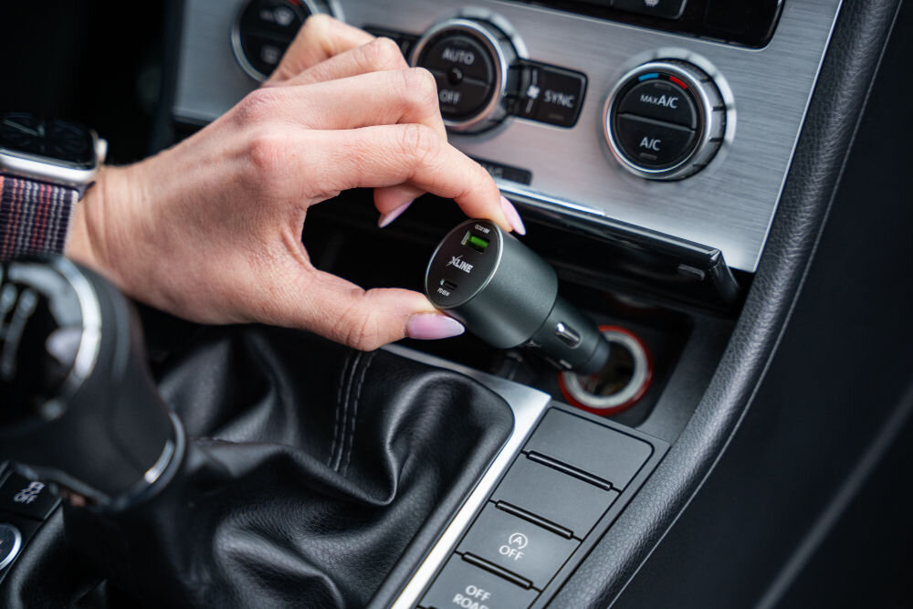 Osoba wkłada ładowarkę samochodową z logo \'XLINE\' do gniazda zapalniczki w samochodzie. W tle widoczne są elementy konsoli samochodowej, w tym pokrętła klimatyzacji.