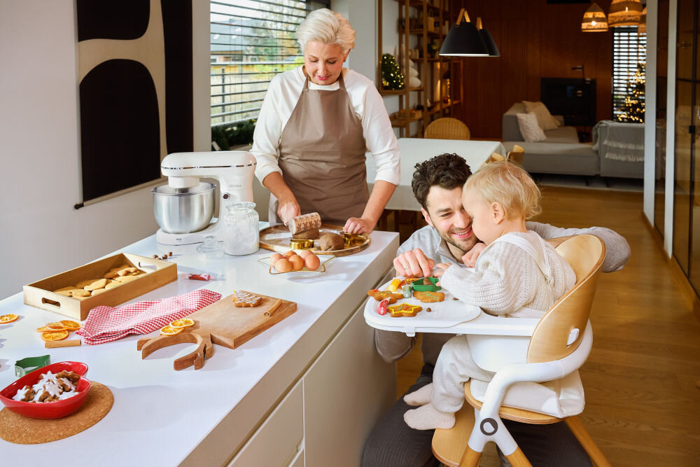 W kuchni znajduje się robot kuchenny planetarny obok składników do pieczenia, takich jak jajka i mąka. W tle widać osobę dorosłą i dziecko w krzesełku, zajętych przygotowywaniem ciastek.