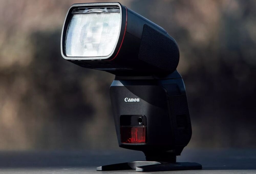Na obrazie widoczna jest lampa błyskowa z logo Canon na ciemnym tle. Lampa ma prostokątny kształt z przezroczystym panelem z przodu.