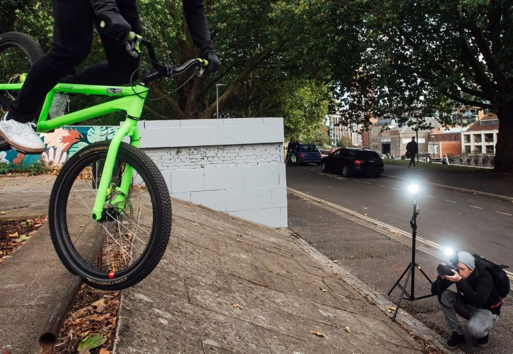 Osoba na rowerze BMX wykonuje trik na rampie, podczas gdy inna osoba fotografuje scenę z użyciem lampy błyskowej na statywie. W tle widać ulicę z zaparkowanymi samochodami i drzewa.