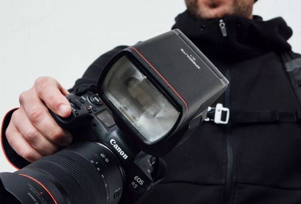 Osoba trzyma aparat fotograficzny z zamontowaną lampą błyskową. Na aparacie widoczne jest logo \'Canon\' oraz napis \'EOS R5\'.