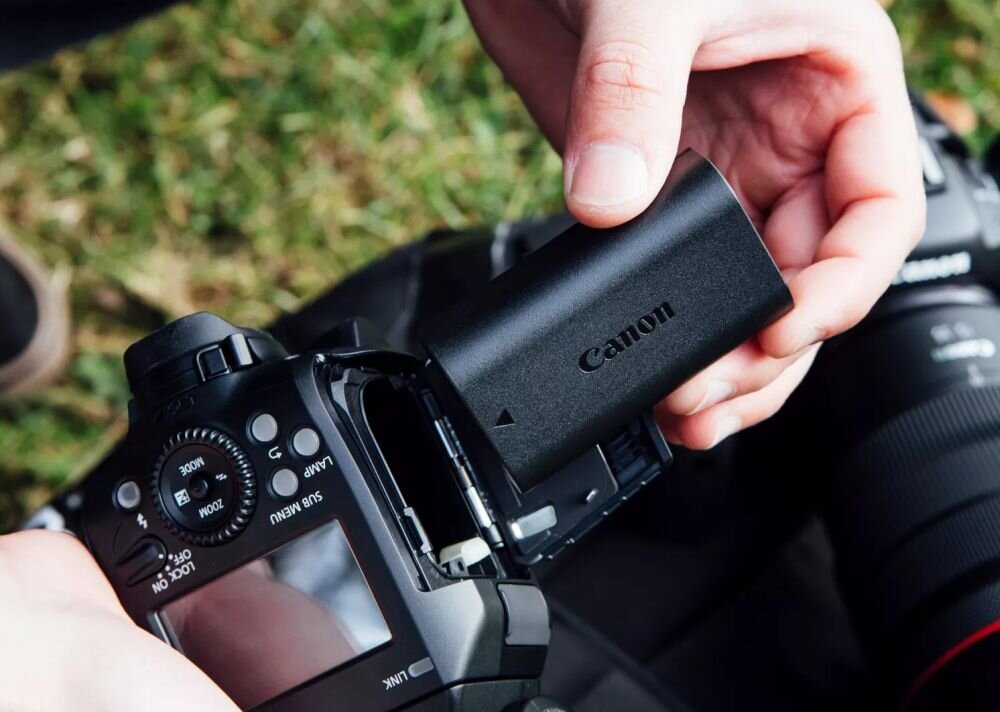 Osoba trzyma czarną baterię z napisem \'Canon\', wkładając ją do aparatu fotograficznego. W tle widoczny jest obiektyw aparatu oraz trawa.