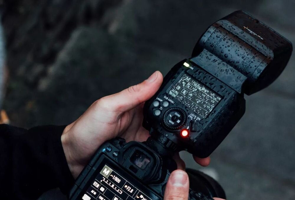 Osoba trzyma aparat fotograficzny z zamontowaną lampą błyskową, na której widoczne są krople wody. Na ekranie lampy widoczny jest tekst z ustawieniami, a obok świeci się czerwona dioda.