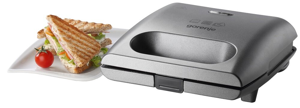 Na obrazie widoczny jest opiekacz w kolorze srebrnym z logo \'gorenje\' na pokrywie. Obok znajduje się talerz z dwoma tostami i pomidorem koktajlowym.
