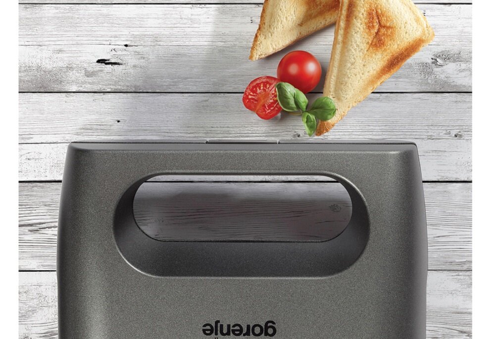 Na drewnianym tle znajduje się opiekacz z widocznym logo \'gorenje\'. Obok leżą dwa trójkątne tosty oraz pomidory i liście bazylii.